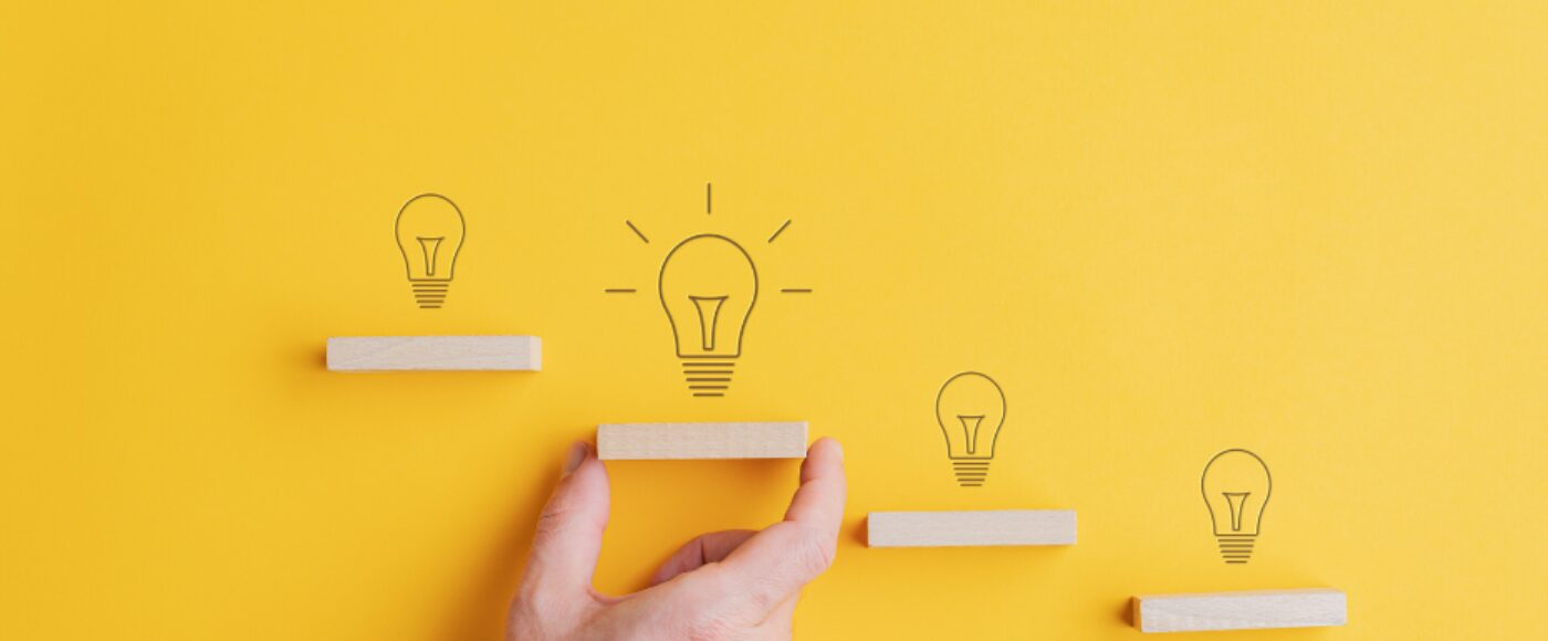 Light bulbs drawn above each step In conceptual stairway of wooden pegs with male hand supporting one with a lit up bulb highlighting innovation